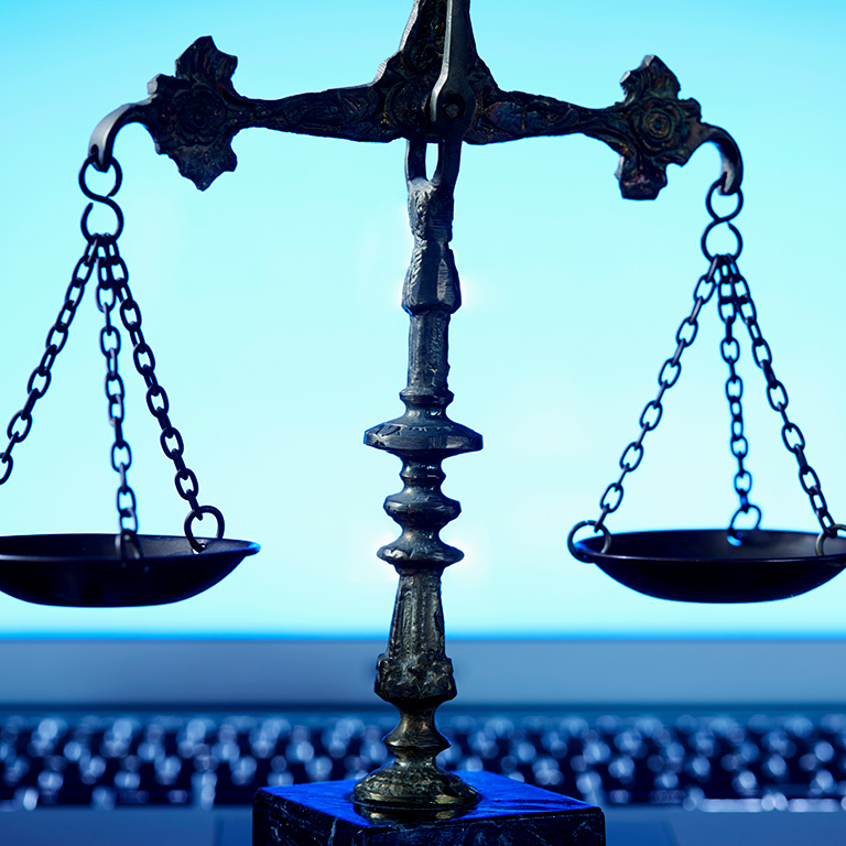 A close-up of a brass balance scale placed in front of an open laptop. The laptop screen glows with a bright blue light, creating a silhouette effect on the scale's chains and pans.