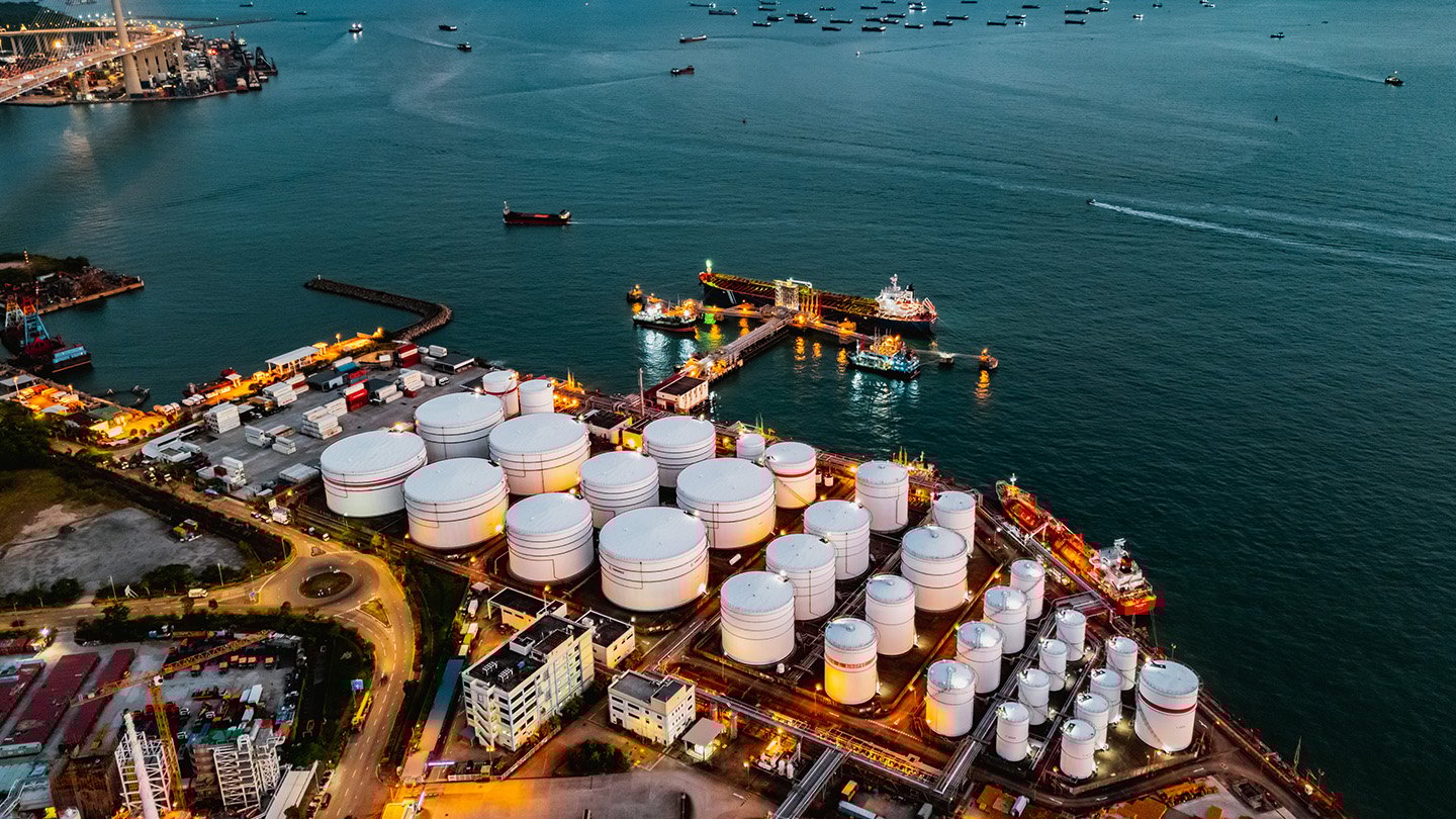Aerial view of an industrial port with large white storage tanks, docked ships, and illuminated facilities near the water. Numerous vessels are visible in the surrounding sea.