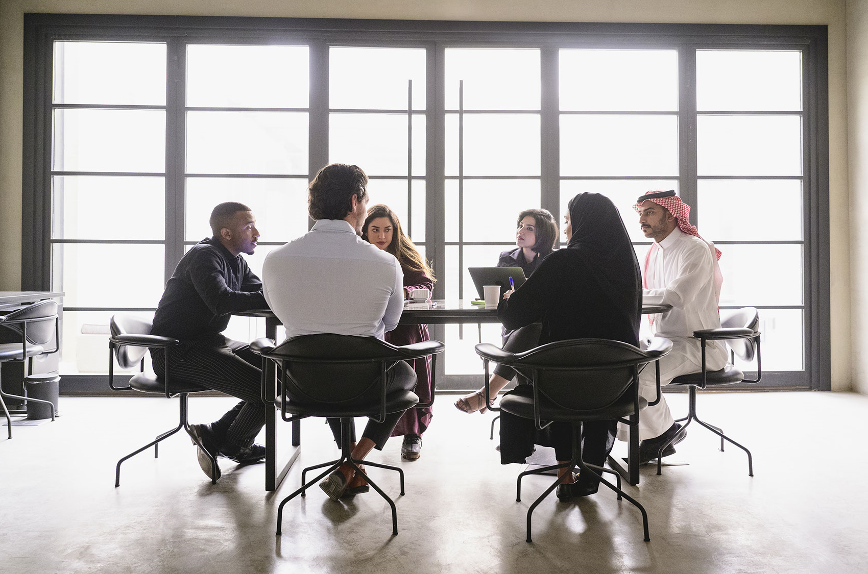 A group of people sitting around a table in a meeting room with large windows in the background. Some are using laptops and taking notes.