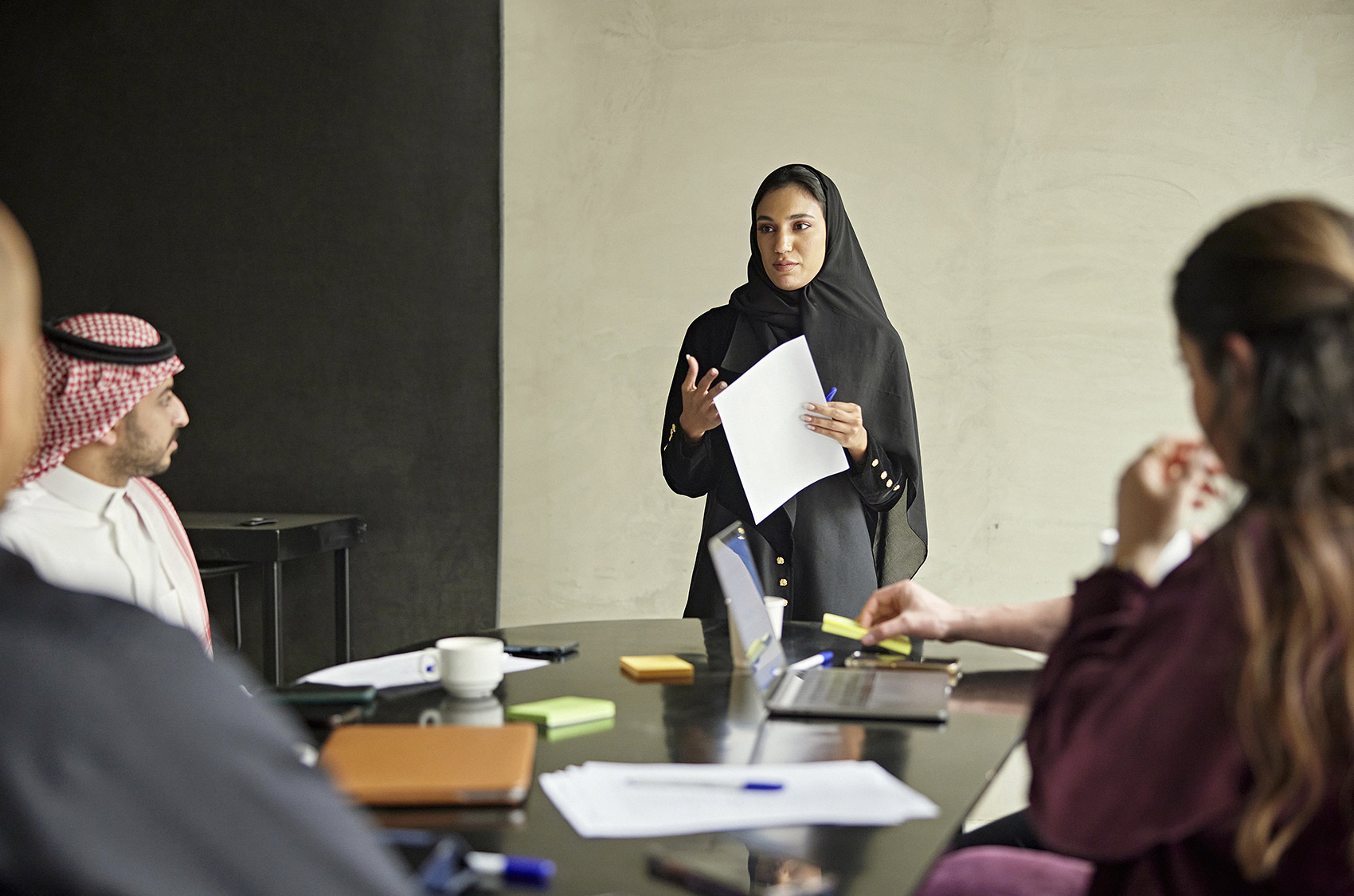 A person in a black outfit is standing and holding papers, speaking to a group of people seated around a table with laptops, notebooks, and coffee cups.