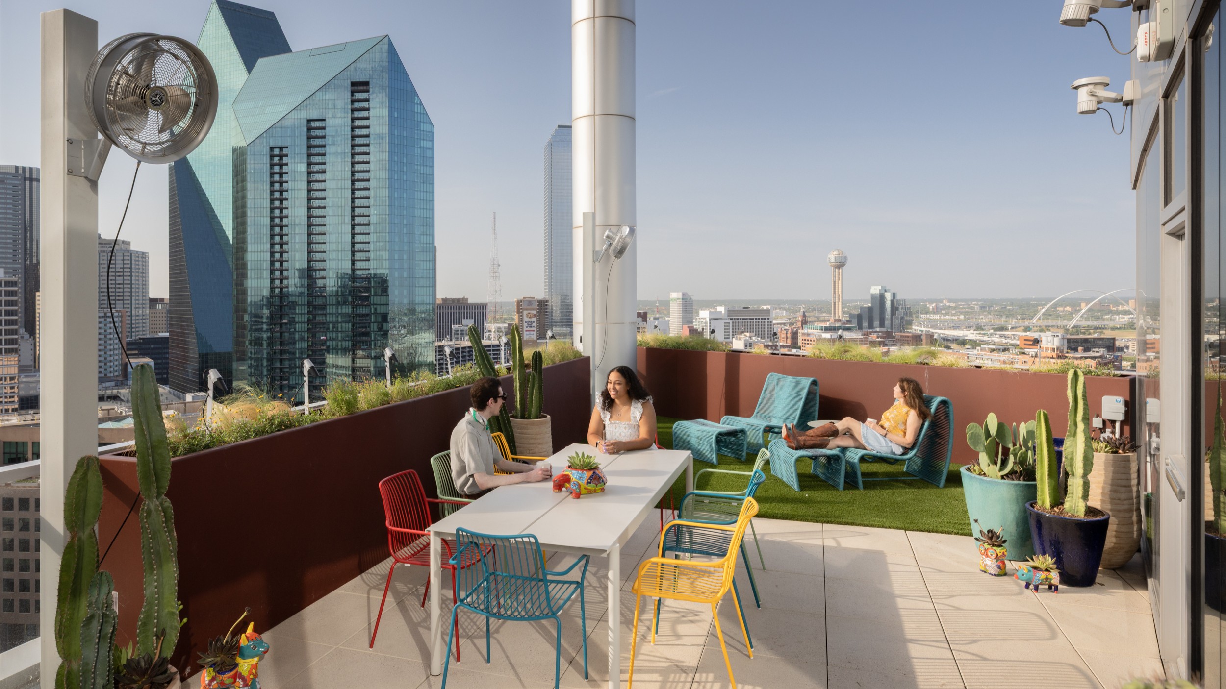 Rooftop patio with three people, colorful chairs, lounge seats, potted cacti, and a city skyline view.