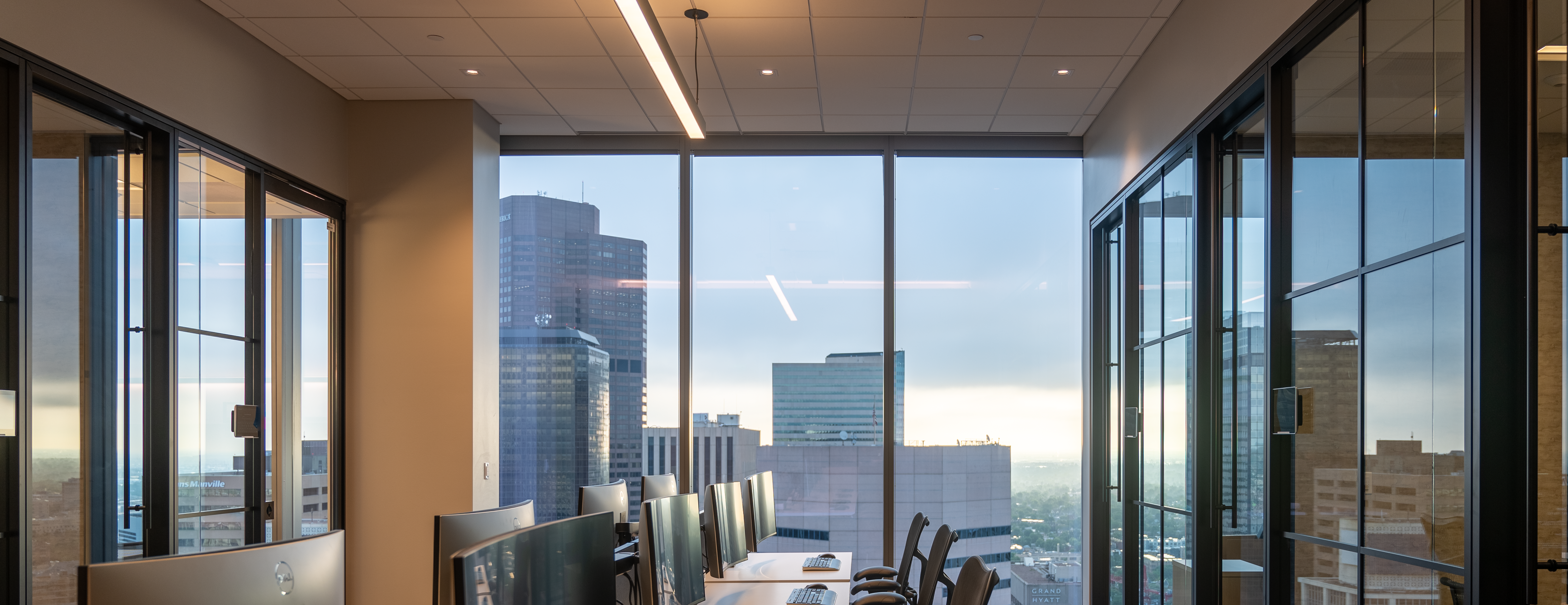Office workspace with desks and monitors beside floor-to-ceiling windows overlooking downtown buildings at dusk.