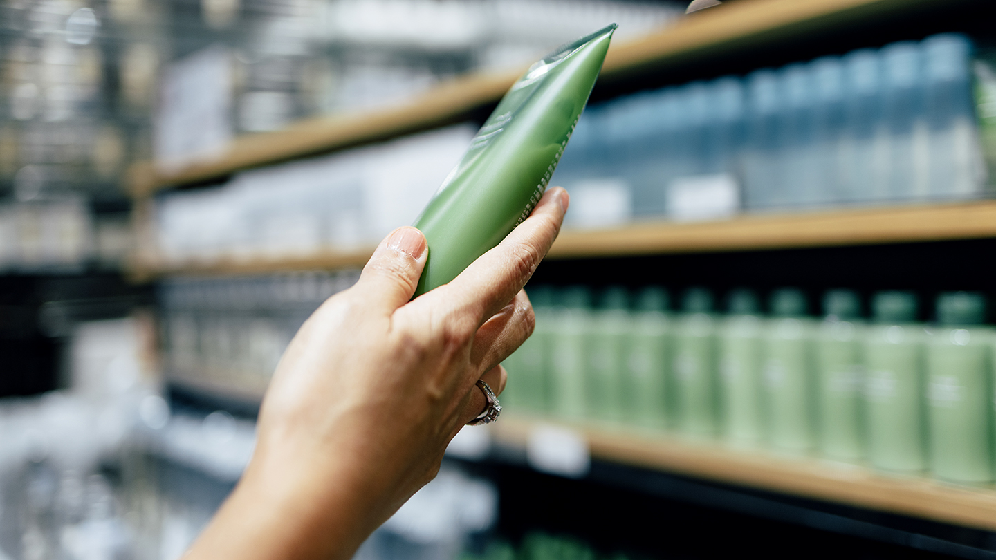 Hand holding a green tube in front of shelves filled with similar green bottles in a store.