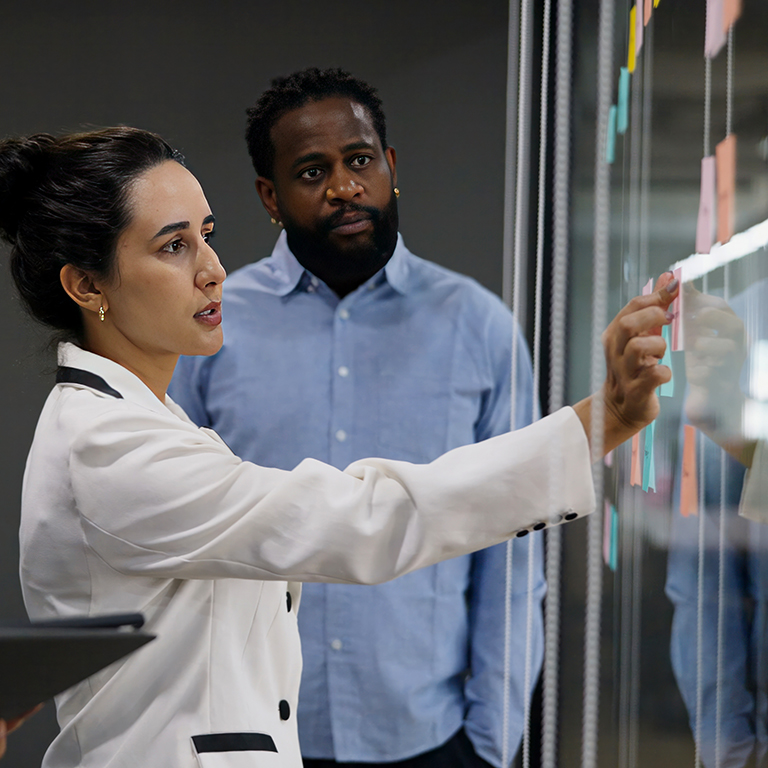 Two individuals are standing in front of a glass wall with sticky notes, one pointing at the notes.