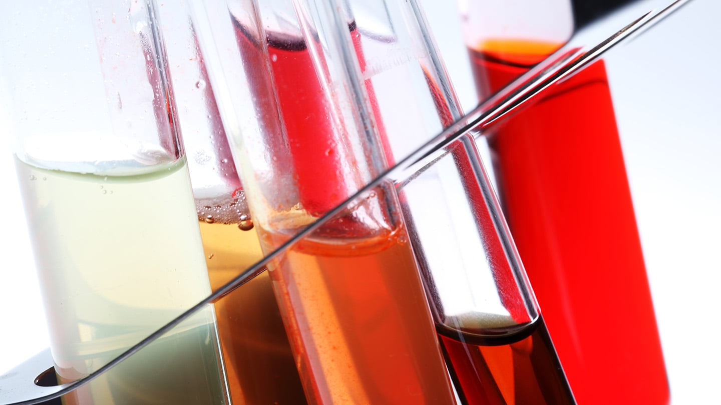Photo of glass test tubes in a metal rack, each filled with a different colored liquid, from pale green to bright red.