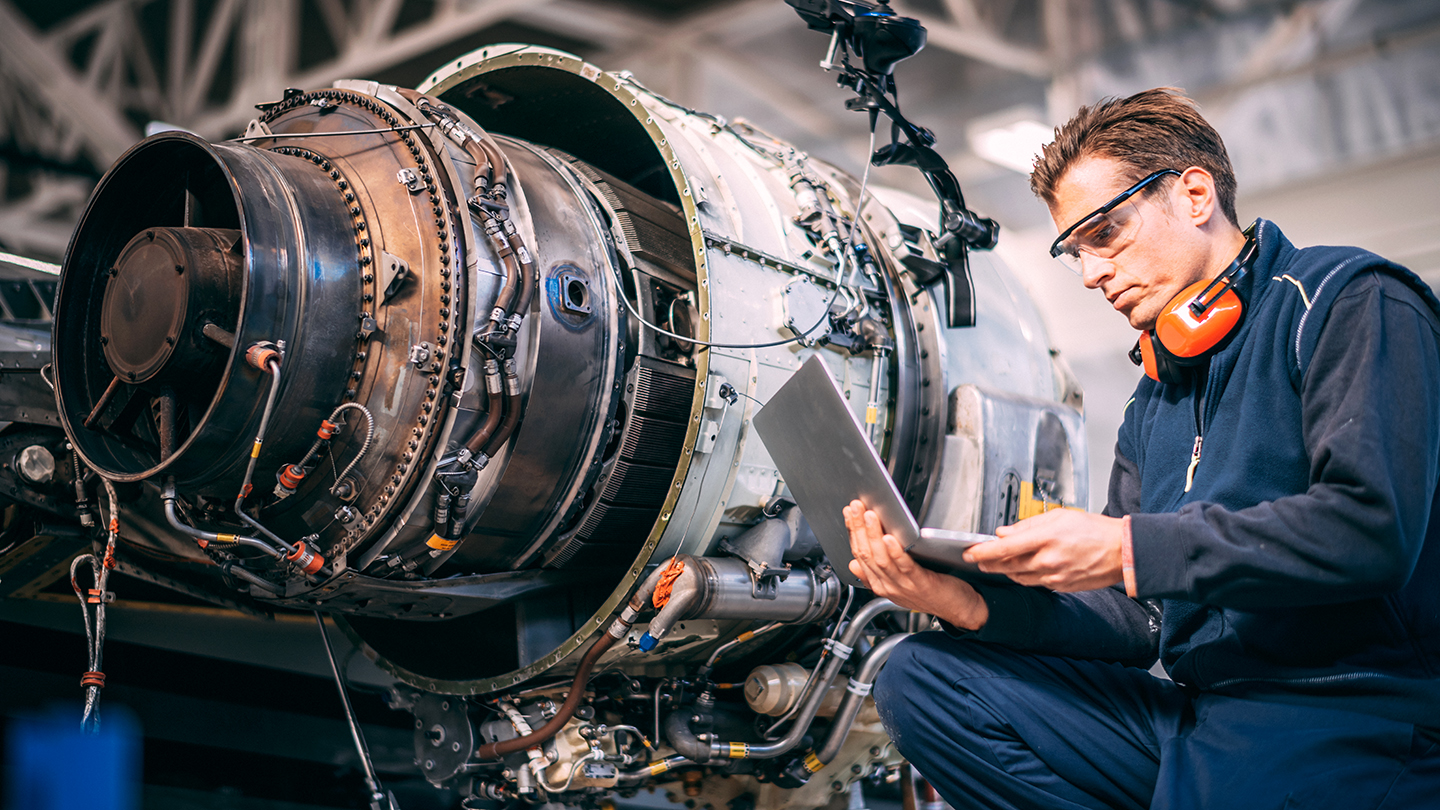 Individual inspecting a jet engine with a laptop, wearing ear protection, in an industrial setting.