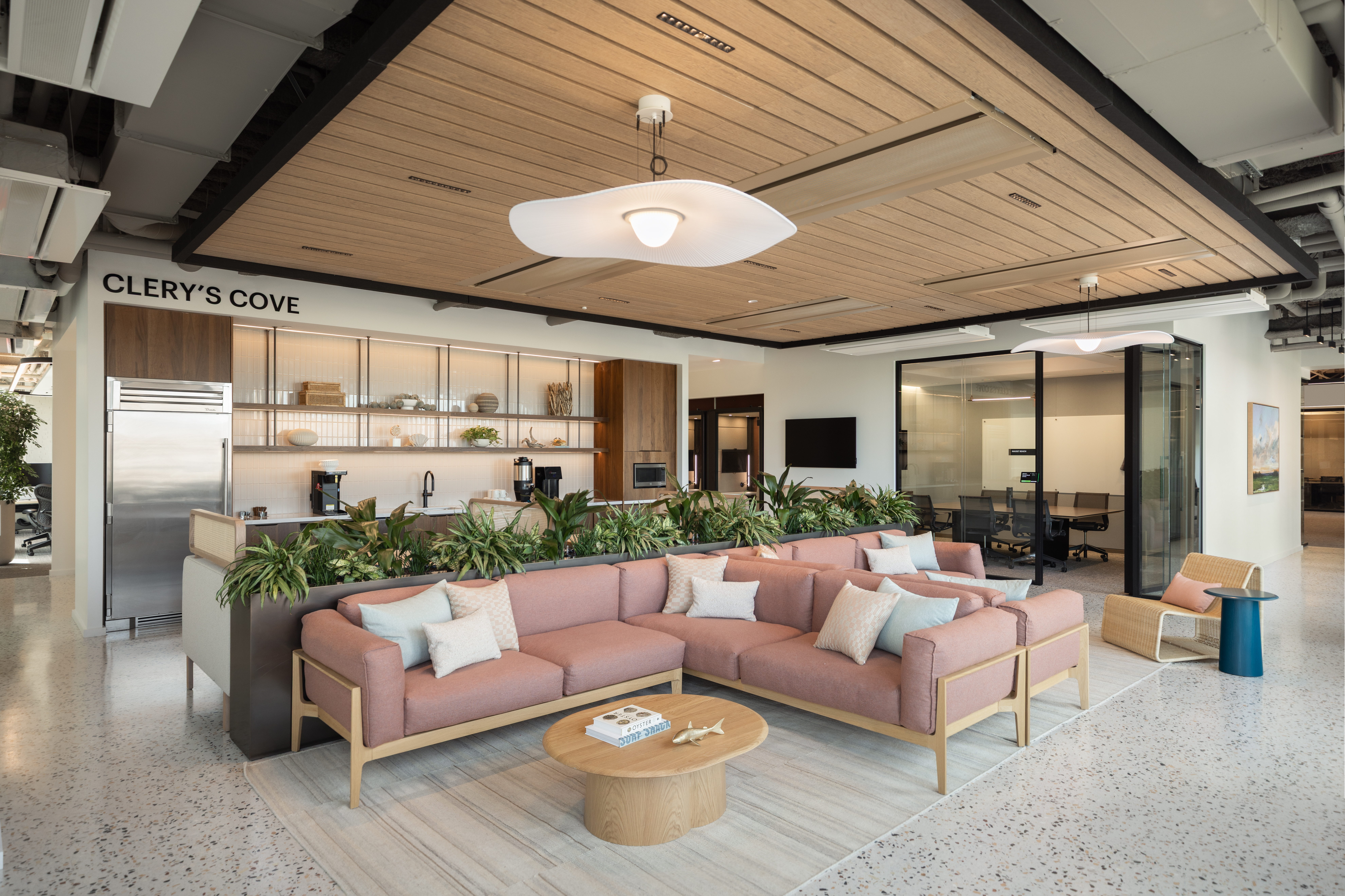 Modern office lounge with pink sectional sofa, round wood table, kitchenette, plants, and glass meeting room under wood-paneled ceiling.