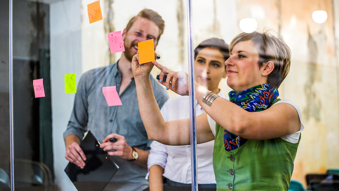 Three individuals collaborating, placing colorful sticky notes on a glass wall. One person holds a tablet.