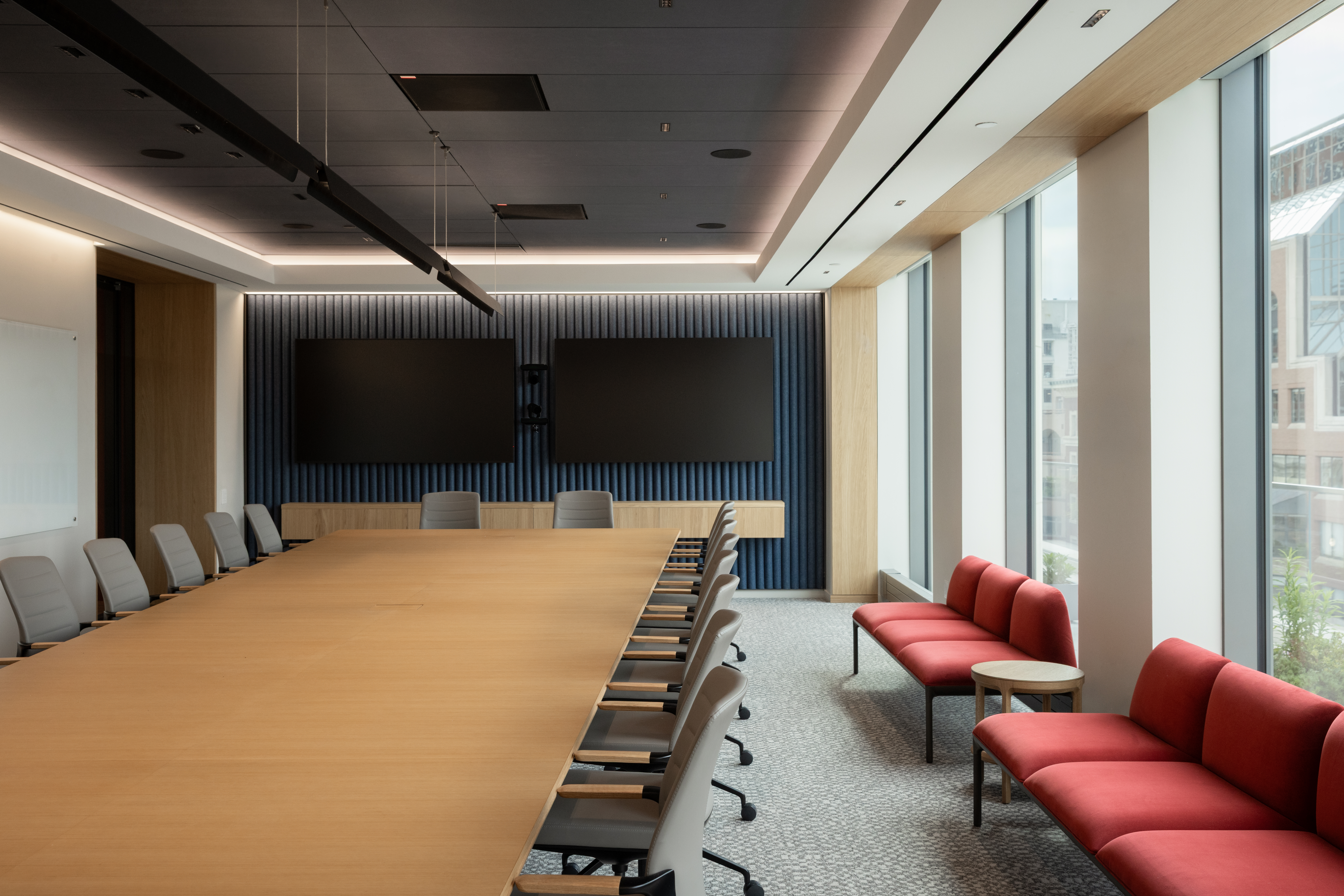 Modern conference room with long wooden table, rolling chairs, two wall-mounted screens, and red lounge seating by tall windows.