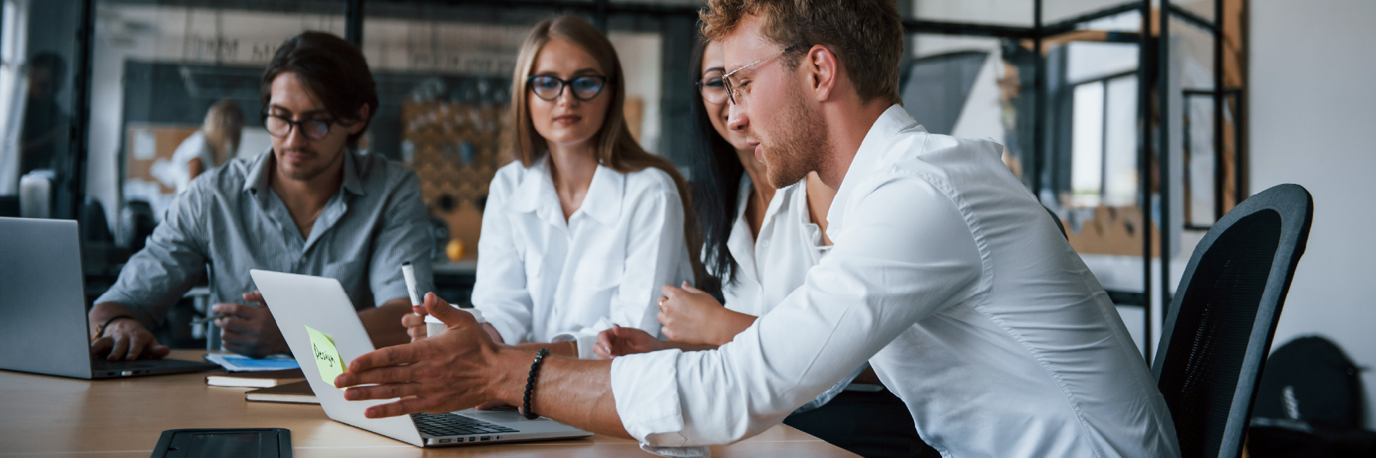 Group of people in a meeting room, discussing around a table with laptops and documents.