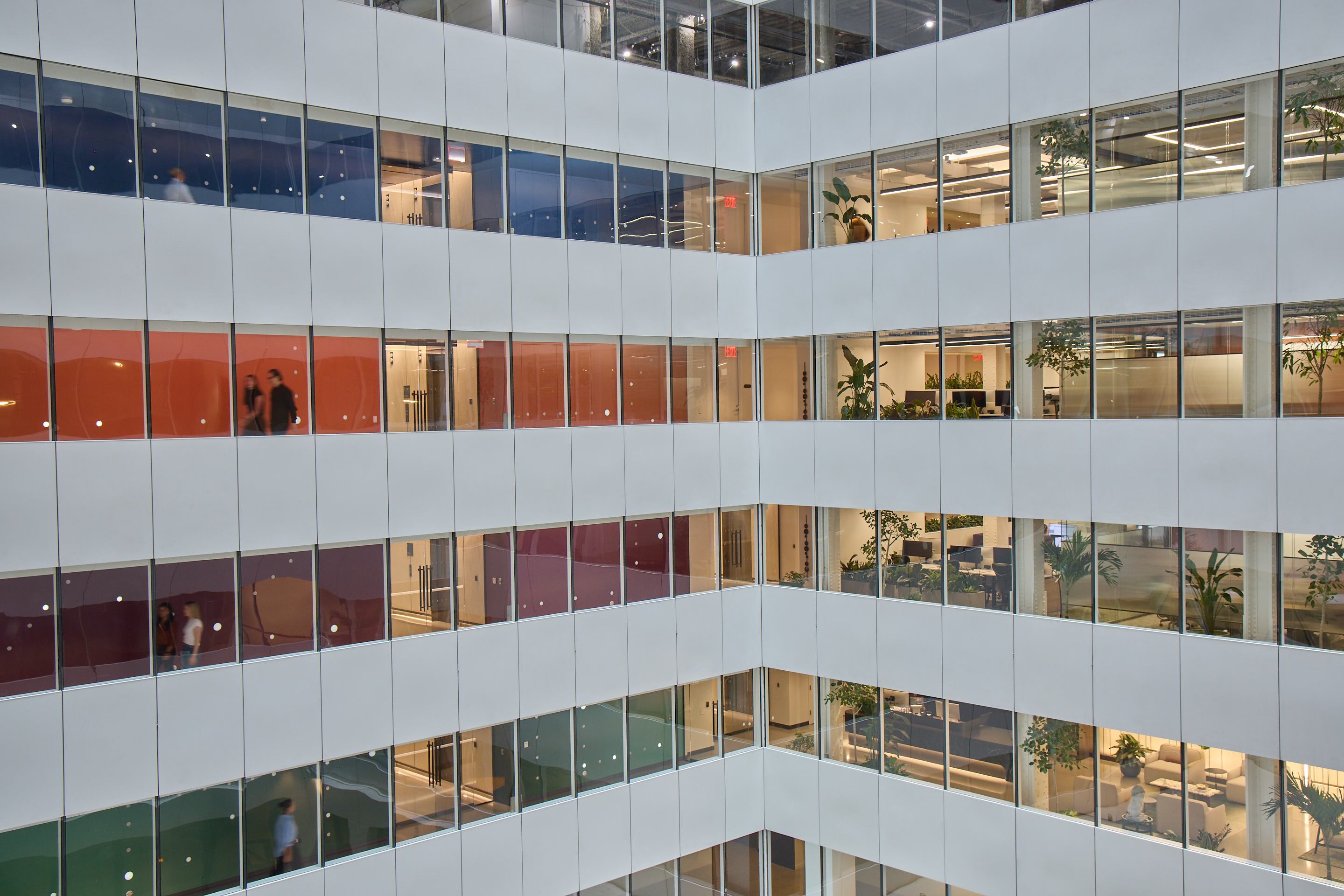 Multi-story office atrium with glass-walled corridors, lit workspaces, plants, and a few people walking on different floors.