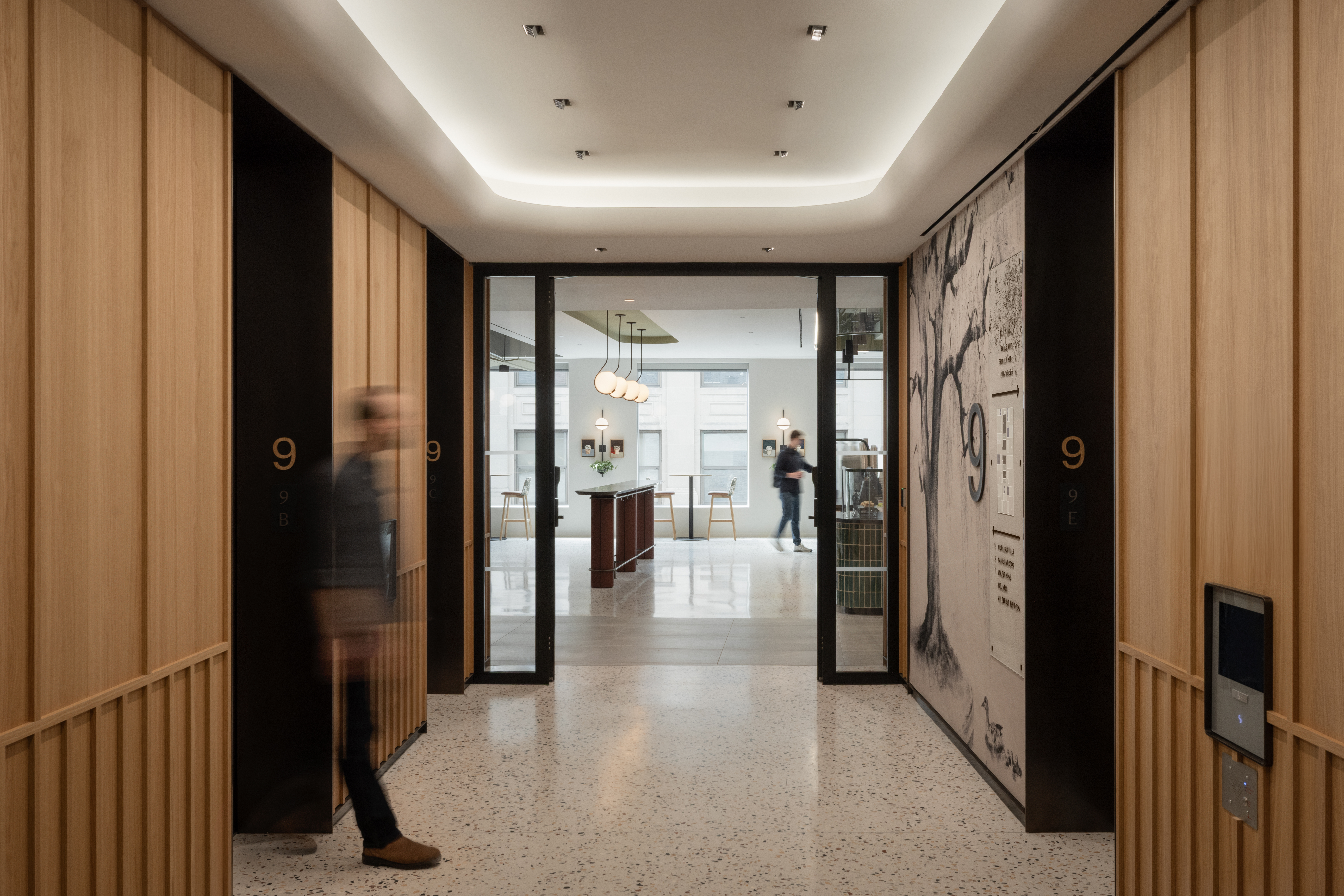 Modern elevator lobby with wood-paneled walls, floor 9 signage, terrazzo floor, and blurred people near a bright hallway.